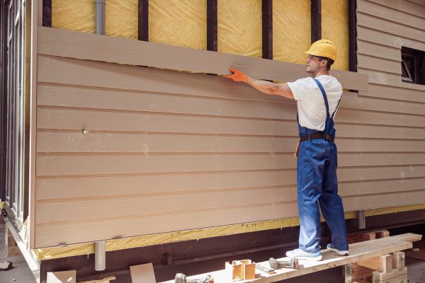 Smiling young man construction worker wearing safety helmet and work overalls while installing exterior wood siding
