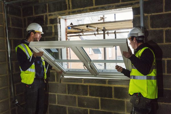 Two builders in hard hats and high visibility jacket fitting a window frame.
